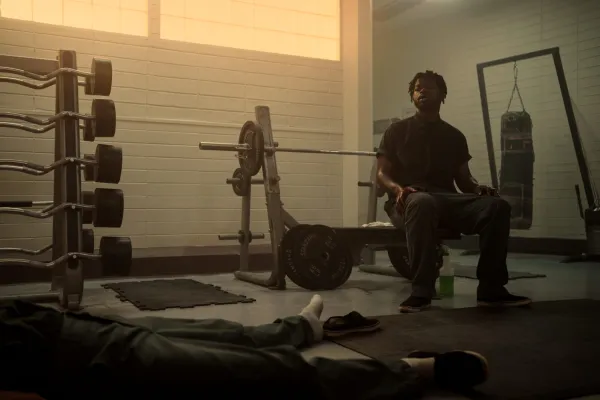 Furly Mac como Christopher Scarver sentado olhando para um cadáver no ginásio de uma prisão.