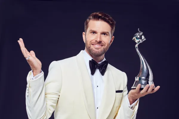 Joel Dommett vestindo um terno branco e segurando um troféu da NTA
