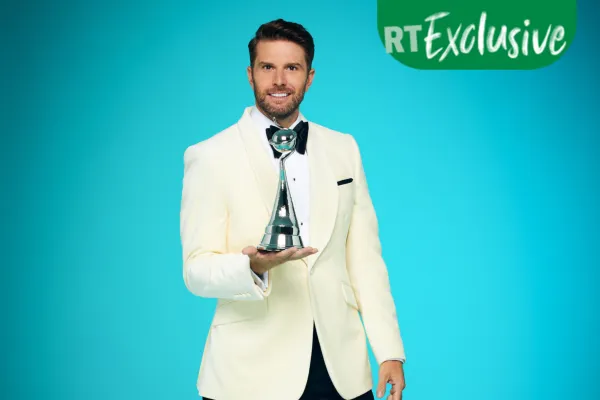 Joel Dommett para o The National Television Awards 2023 vestindo um terno branco, segurando um troféu NTA, com RT Exclusive no canto superior direito