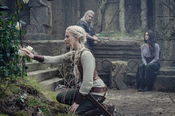 Ciri olhando para uma flor, com Geralt e Yennefer encostados ao fundo
