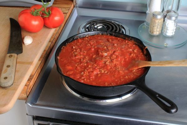 Сос за спагети от домати и месо, задушаващ в чугунен тиган върху електрическа печка от неръждаема стомана с домати и чесън върху дъска за рязане с голям кухненски нож.
