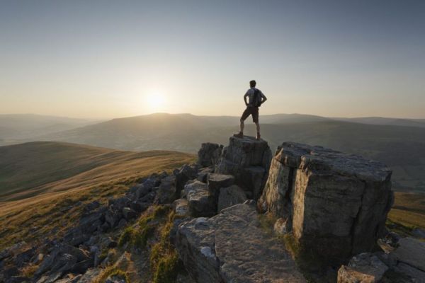 vyras stovi ant kalno viršūnės su saulėlydžiu