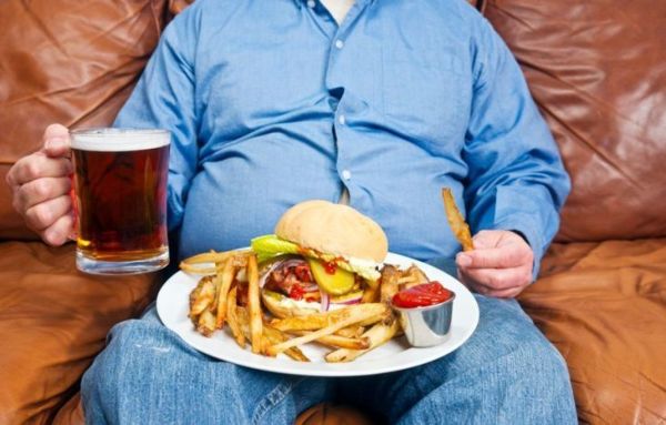 صورة لرجل بدين يجلس على أريكة قديمة مع وجبة كبيرة غير صحية على حجره ونصف لتر من الجعة في يده. السمنة هي سبب رئيسي لمرض السكري.